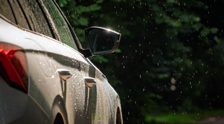 A silver car driving through falling rain with greenery behind